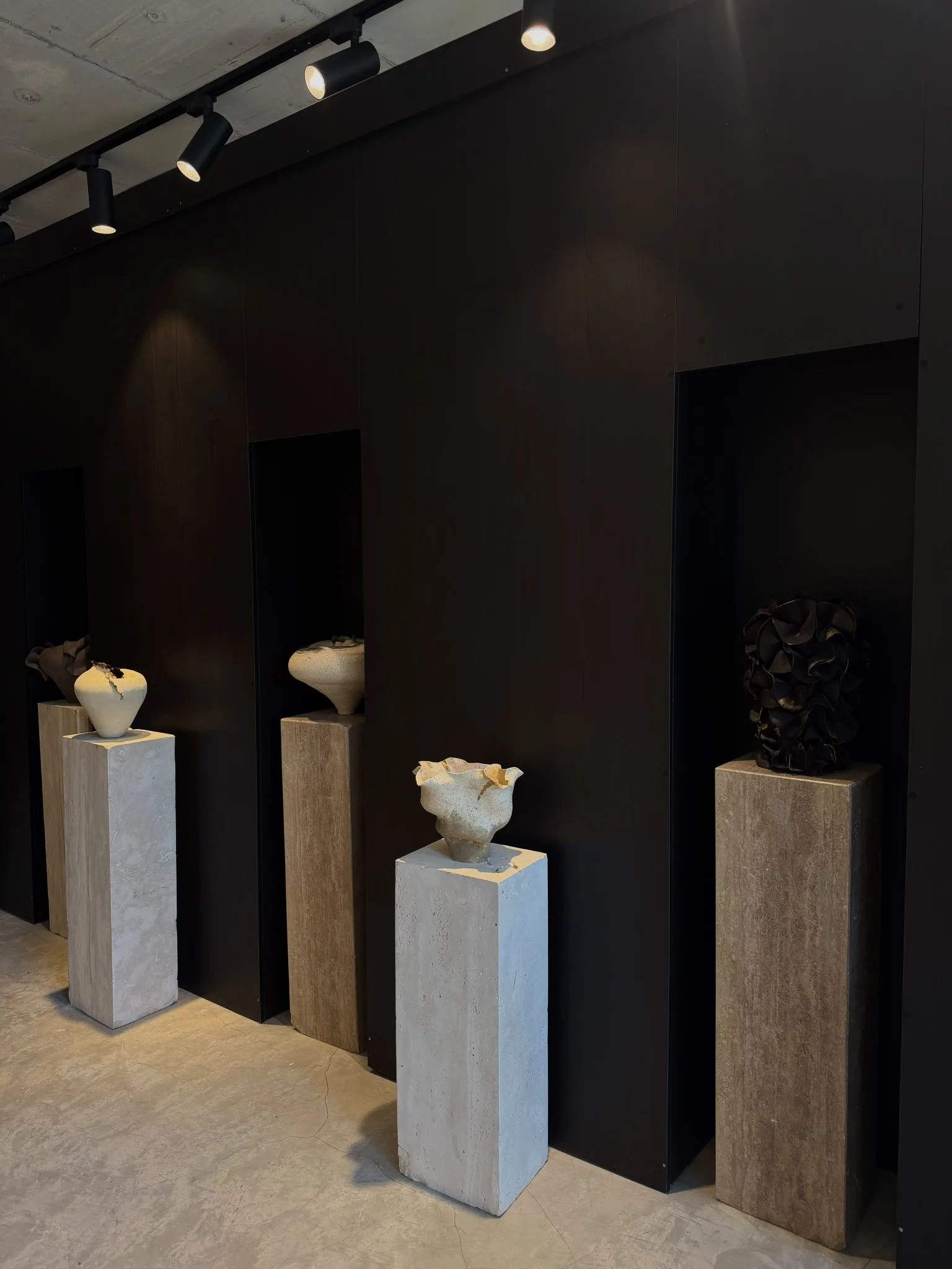 Travertine plinths lined along a black wall, carrying pale and dark ceramics